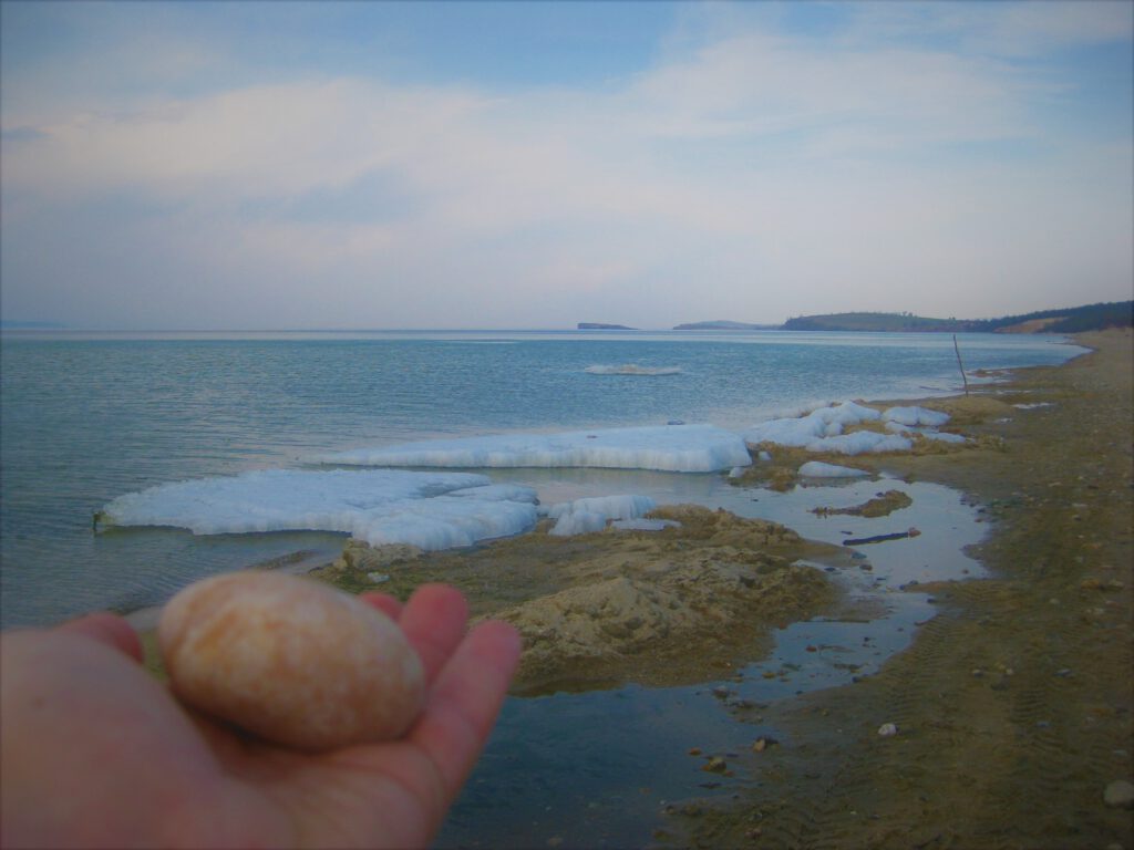 Blick über den blauen Baikalsee mit restlichen Eisschollen am Ufer. Eiförmiger Stein in der Hand, links unten im Bild. 