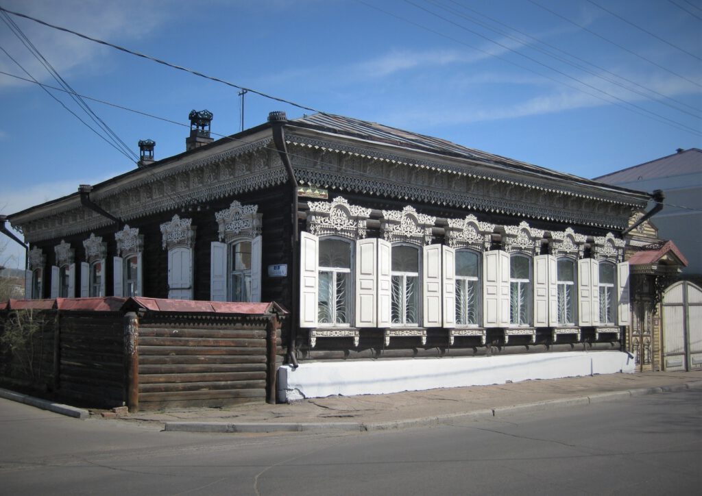 Sibirisches Holzhaus in Ulan Ude an einer Straßenecke. Fenster haben weiße Fensterklappen. 