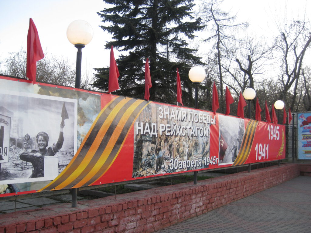 "Banner des Sieges über den Reichstag" an einem Zaun in Krasnojarsk. Darauf stecken rote Fahnen. 