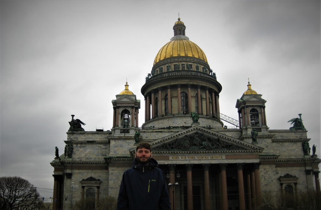 Sehenswürdgkeiten von St. Petersburg. Selfie vor der Isaakskathedrale bei bewölktem Himmel. Massive, kantige Fassade. Darauf zwei kleine und ein Haupt-Turm. Jeweils mit goldener Kuppel. 