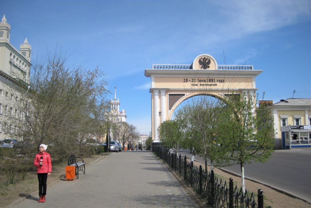 Blick hoch zum Zarentor in der Ulica Lenina in Ulan-Ude. Links steht ein Kind mit Mikrofon auf dem Bürgersteig. 
Царские ворота