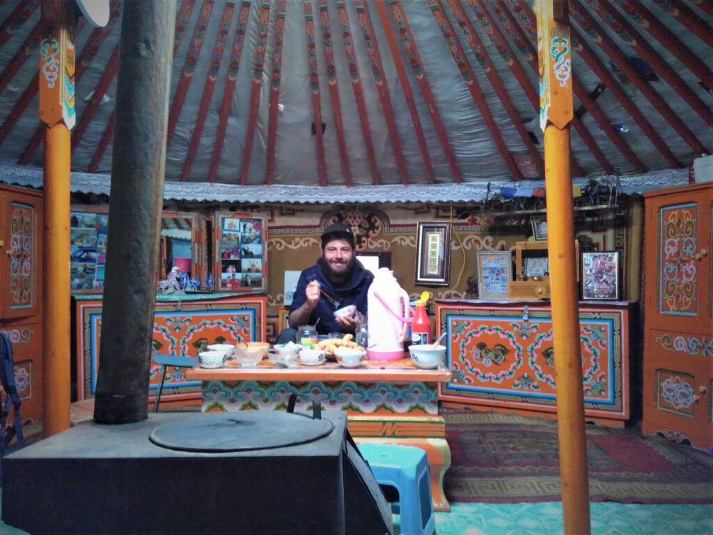 Ich sitze beim Essen am Tisch in einem mongolischen Ger. Im Vordergrund der Ofen. Im Hintergrund bunte Möbel. 