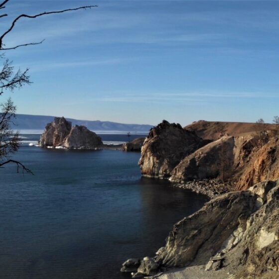 Blick auf den Schamanenfelsen über das Blaue Wasser des Baikalsees. Links im Bild ein kahler Baum. Rechts die steile Küste Olchons. Blauer Himmel.