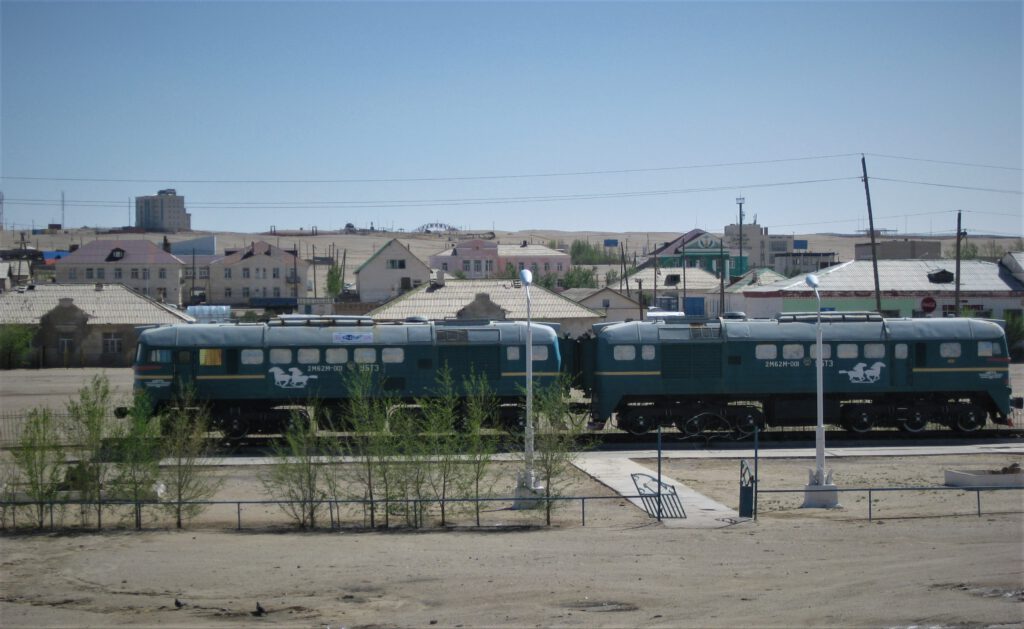 Mongolische Triebwagen in grüner Farbe mit weißen Pferdesymbolen stehen auf einem Abstellgleis. Im Hintergrund eine kleine Wüstgenstadt.