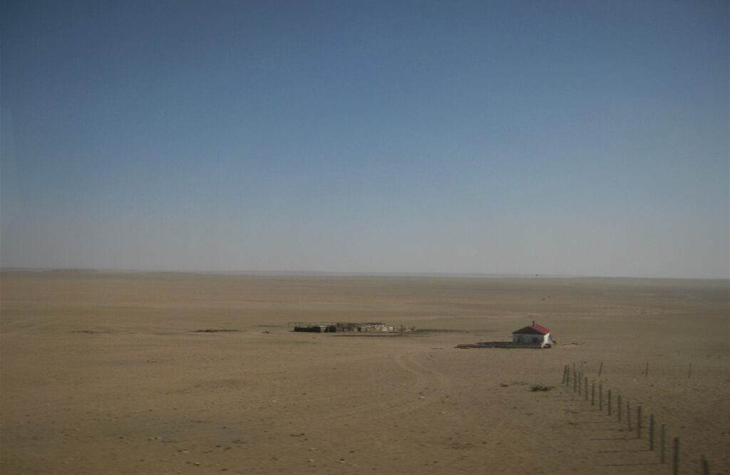 Mit dem Zug durch die Wüste Gobi. Ein kleines Haus, mitten in der Wüste. Daneben vermutlich ein Gehege. 