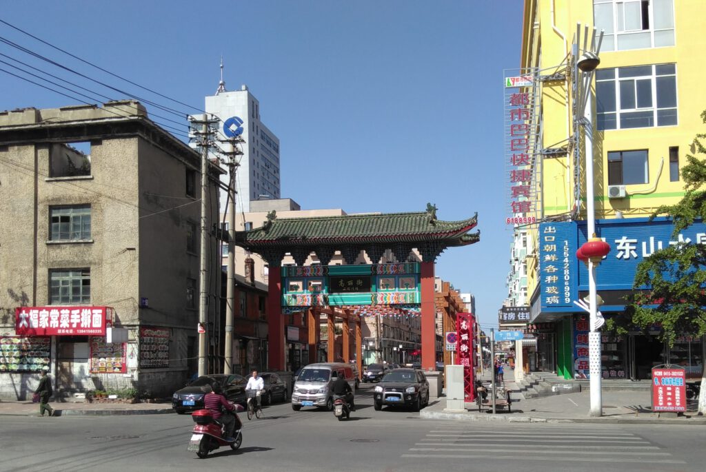 Straßenaufnahme des Eingangs zur Koryo Street in Dandong. Autos fahren durch ein traditionelles chinesisches Tor. 