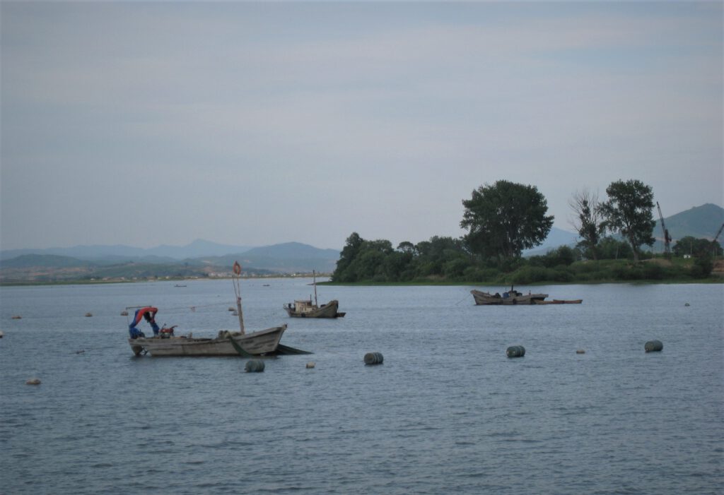 Dandong: Drei keine, graue Fischerboote liegen auf dem Yalu-Fluss vor der nordkoreanischen Küste. Im Hintergrund die hügelige Landschaft Nordkoreas.
