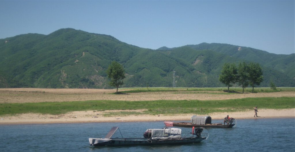 Zwei Fischerboote auf dem Yalu, Ein nordkoreanischer Soldat spaziert mit Gewehr am Ufer von den Booten weg.