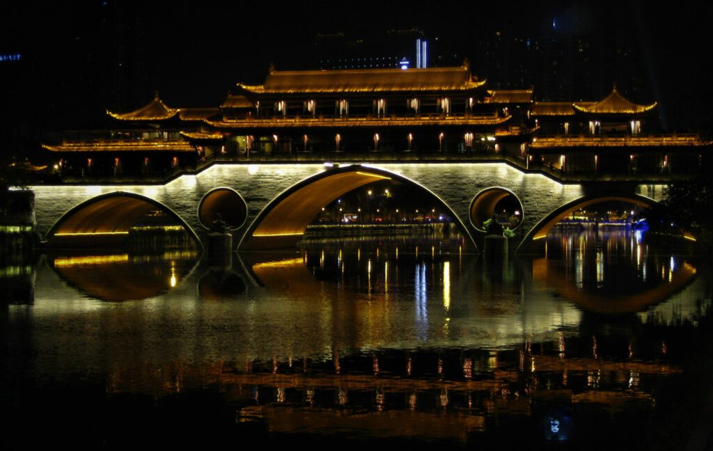 Beleuchtete Anshun Brücke bei Nacht. Das Licht spiegelt sich verschwommen im Wasser. 