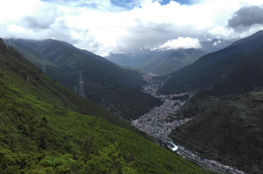 Blick hinab ins Tal der Stadt Kangding. Die grünen Berge um Kangding mit Strommasten und starker Bewölkung. 