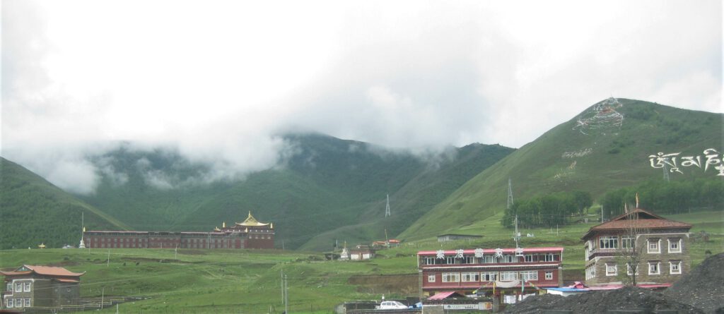 Grüne Hügel in Sichuan. Ein tibetisches Kloster mit goldenem Dach und Wohnhäuser betten sich in die Landschaft ein. Ein Mantra ist auf den Hügel geschrieben. 