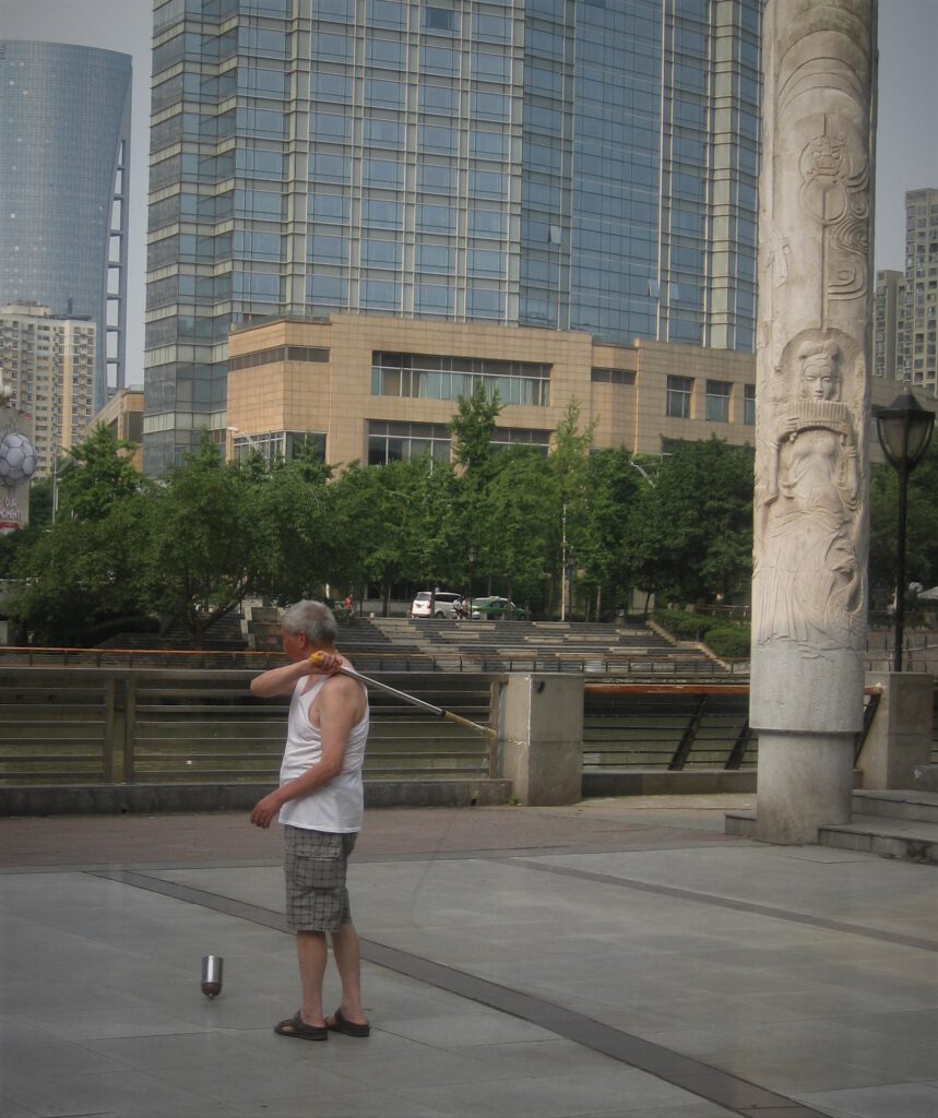 Ein Mann in weißem Unterhemd spielt das Peitschen-Kreisel-Spiel auf einem Platz in Chengdu.