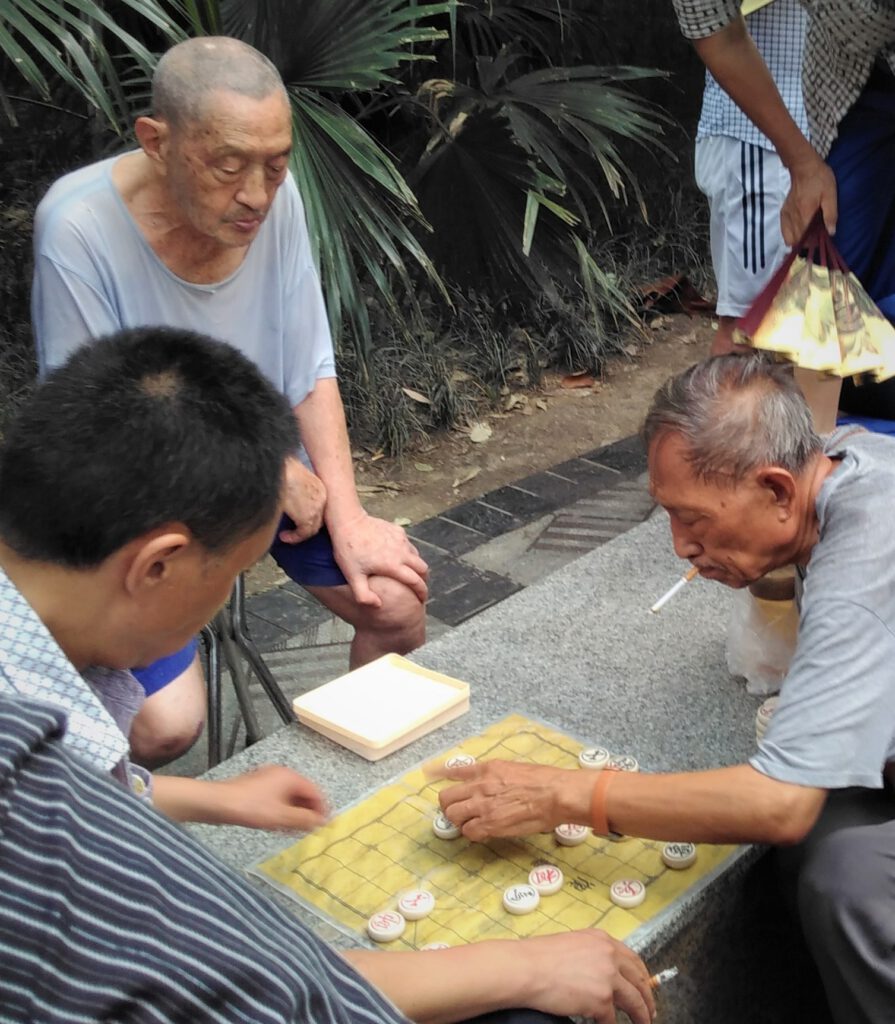 Männer im Volkspark von Chengdu spielen konzentriert Xiangqi, das chinesische Schachspiel. Einer der Männer hat eine Zigarette im Mund und bewegt einen Spielstein. 