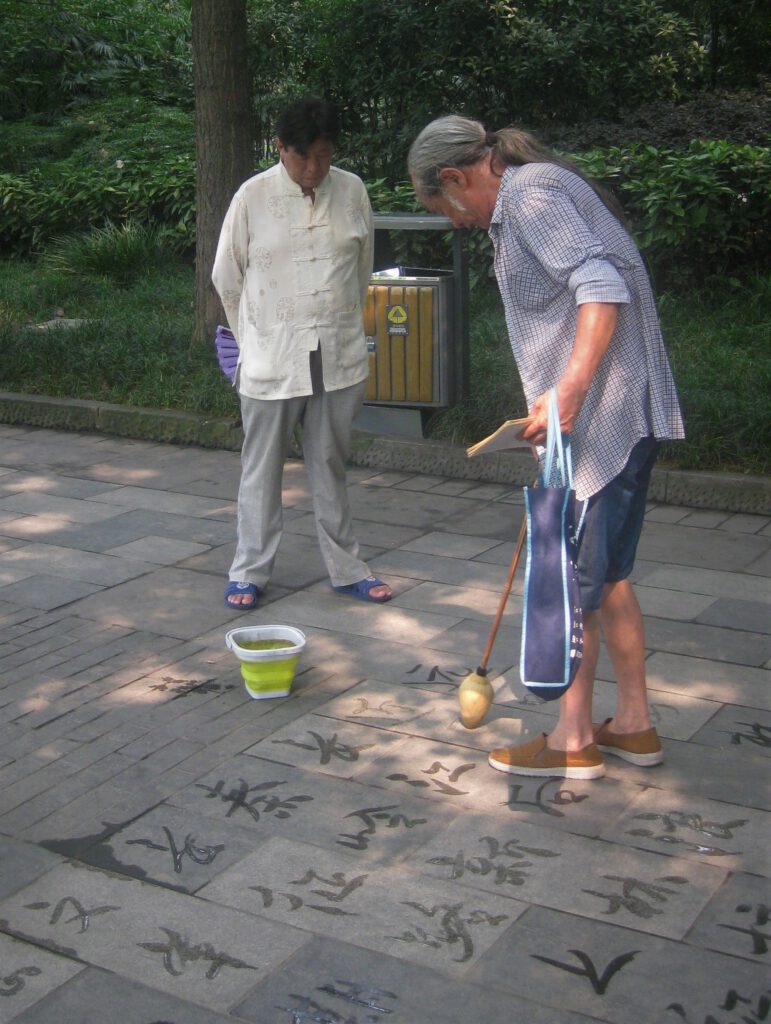Ein Dishu-Künstler zeichnet mit einem großen Pinsel und Wasser chinesische Schriftzeichen auf den Betonboden. Ein Mann sieht ihm zu, 