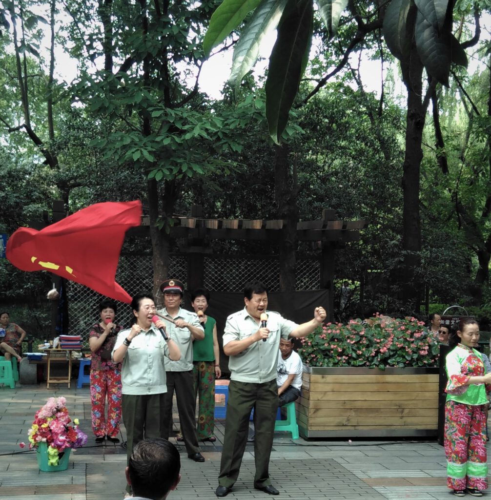 Sängerin und Sänger singen mit geballter Faust im Volkspark von Chengdu. Dahinter ein Mann, der die Fahne der kommunistischen Partei schwenkt.  
