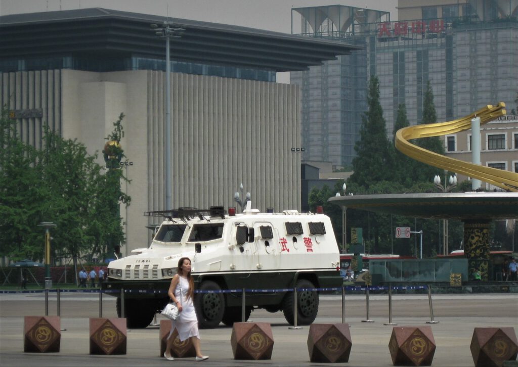 Eine Frau läuft vor einem gepanzerten Polizeiwagen, der auf dem Tianfu-Platz in Chengdu steht. Im Hintergrund eine der beiden Fontänen-Säulen. 