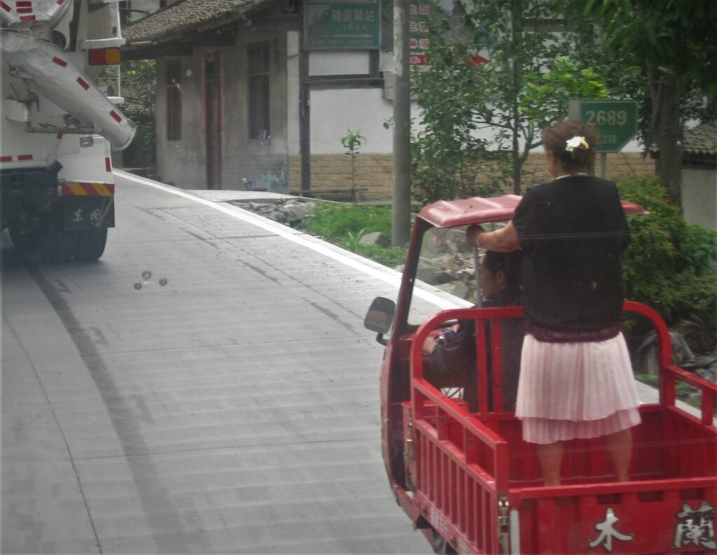 Eine Frau steht auf der Ladefläche eines  kleinen Fahrzeugs. Davor fährt ein Betonmischer. 