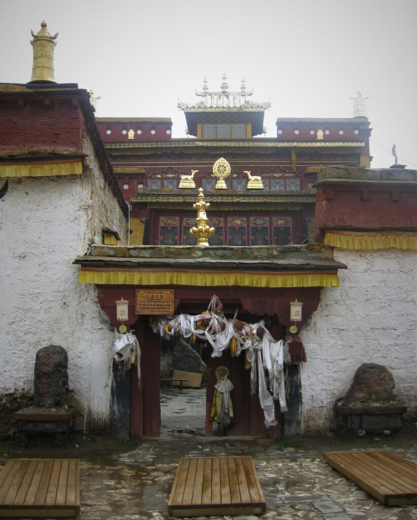 Eingang zum Geburtshaus des 7. Dalai Lama. Weiße Bänder schmücken den Eingangsbogen. Auf den Dächern befinden sich goldene Mandalas und ähnliche Verzierungen. 