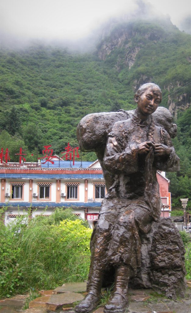 Weibliche Bronzestatue mit geflochtenem Zopf, auf einem Bronzefels sitzend. Im Hintergrund ein grüner Hügel und ein Haus mit chinesischen Schriftzeichen. 
