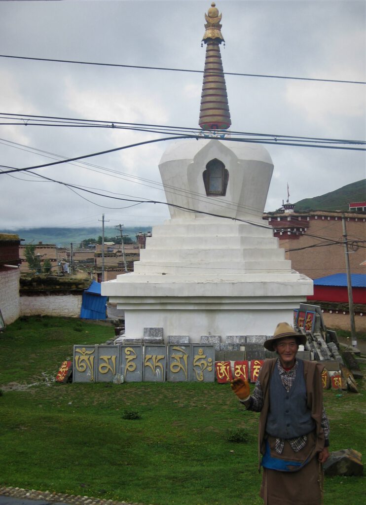 Ein Mann winkt vor einer weißen Stupa. Kacheln mit der Aufschrift "Om mani padme hum" in tibetischer Schrift. 