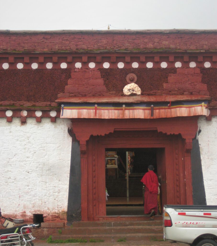 Mönch in roter Robe steigt über die Türschwelle des roten Eingangstors ins Litang Kloster. 