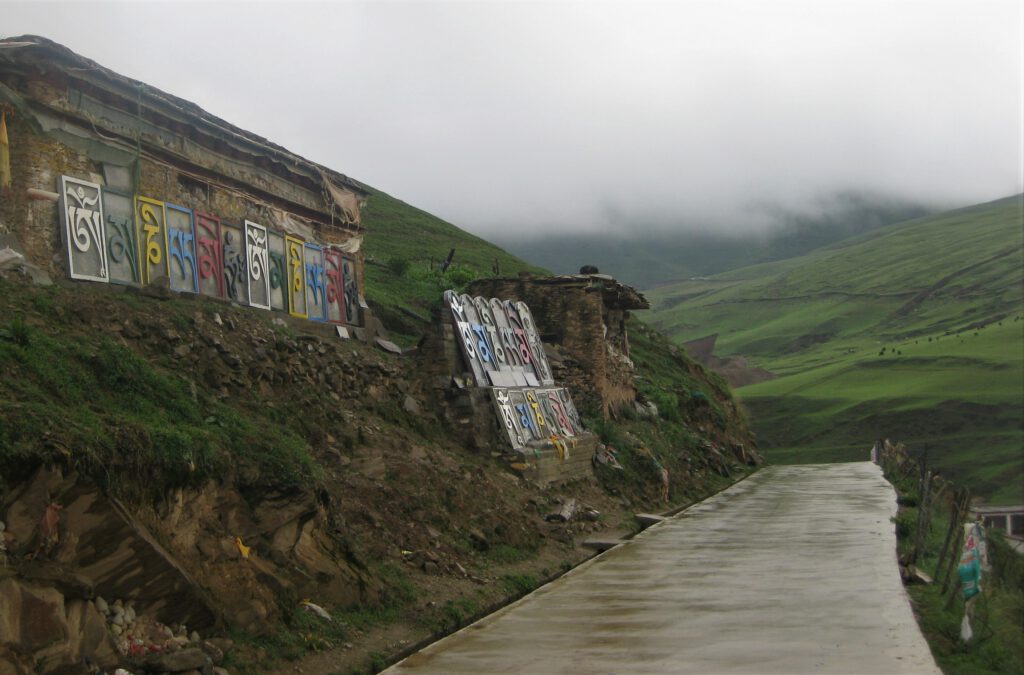 Bunte Kacheln mit der Aufschrift "Om mani padme hum" in tibetischer Schrift schmücken den Rundweg am Litang Kloster. Im Hintergrund die vernebelten, grünen Hügel. 