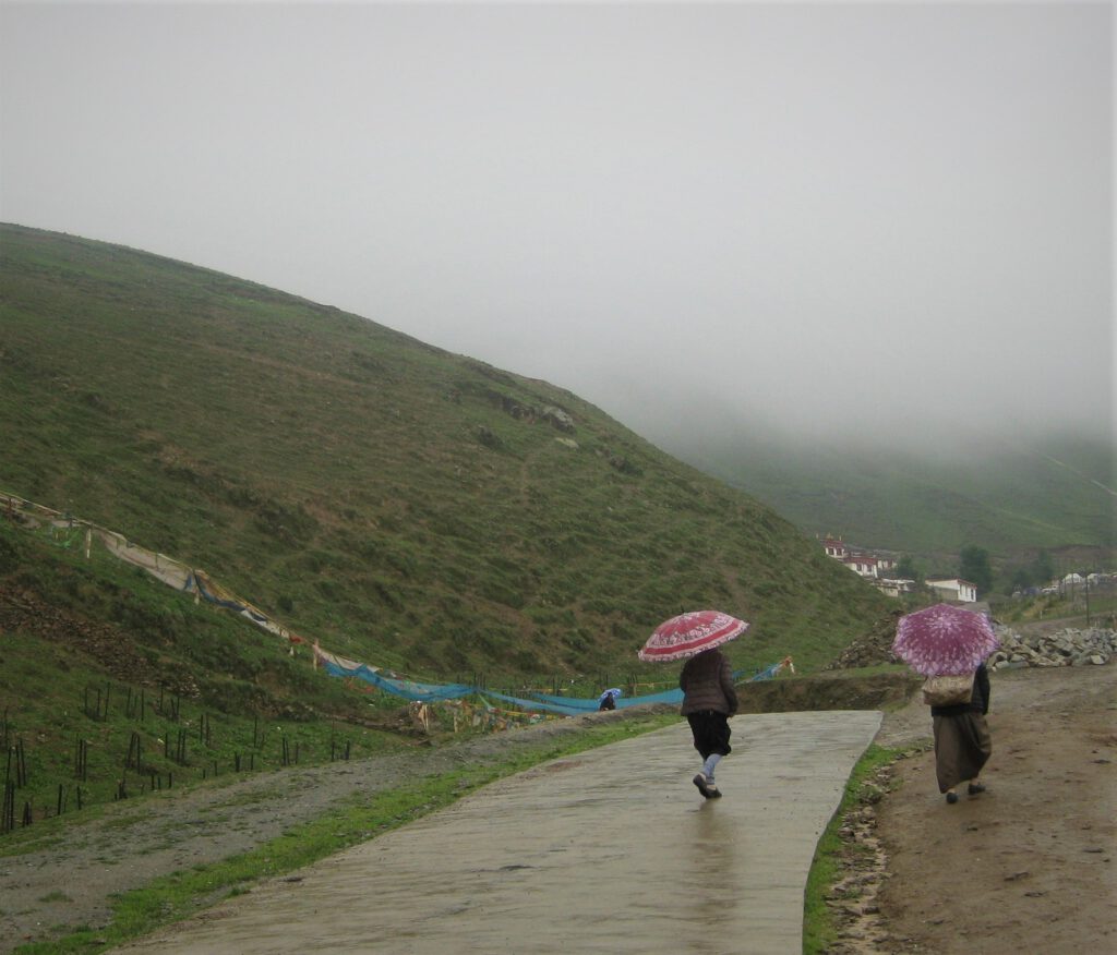 Zwei Frauen mit Regenschirmen auf einem Weg zwischen den vernebelten, grünen Hügeln von Litang. 
