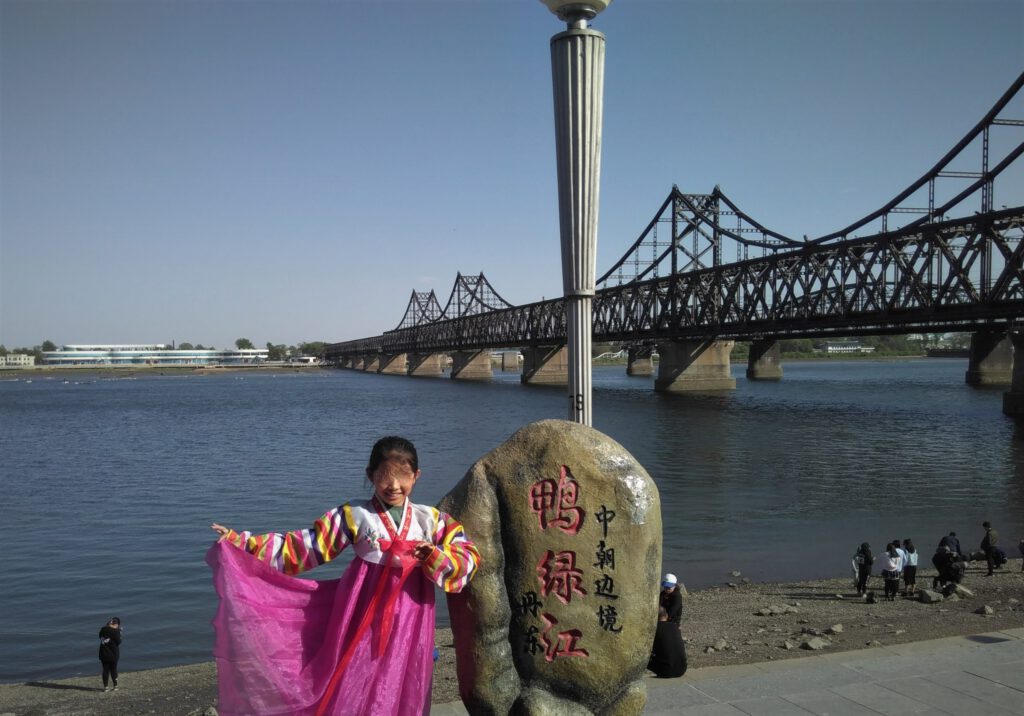 Ein Mädchen in traditionellem, pinkfarbene koreanischen Kleid posiert vor einem Grenzstein vor dem Yalu-Fluss mit der Freundschaftsbrücke. 