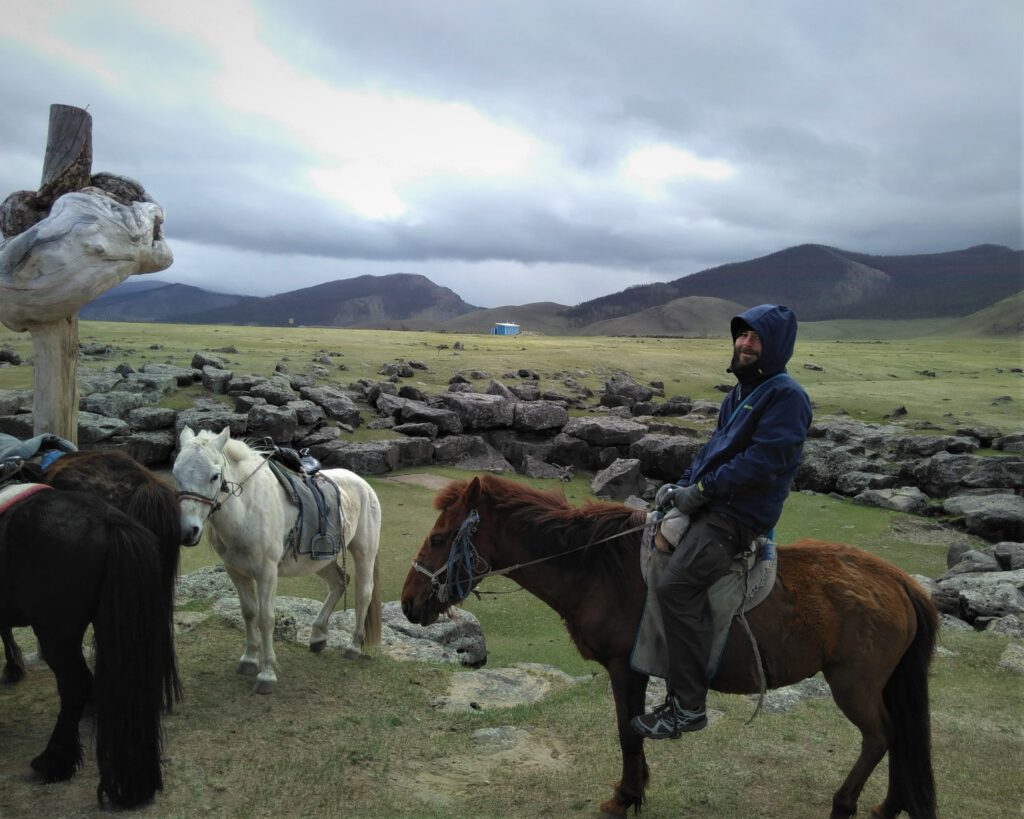 Ich sitze auf einem mongolischen Pferd. Daneben sind weitere Pferde an einem Holzpfahl angebunden. Im Hintergrund das bewölkte Orchon-Tal.
