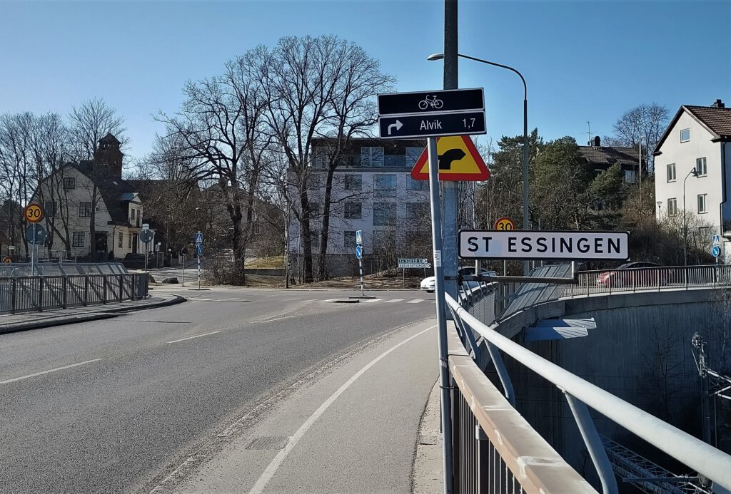 Ortseingang nach Stora Essingen auf der Essingebron. Auf einem weißen Schild steht "St Essingen". Die Straße mündet in eine Kreuzung.