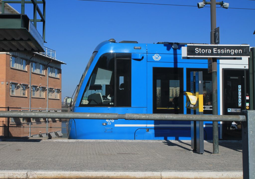 Blaue Straßenbahn der Tvärnabanan steht an der Haltestelle Stora Essingen. Seitenansicht. 