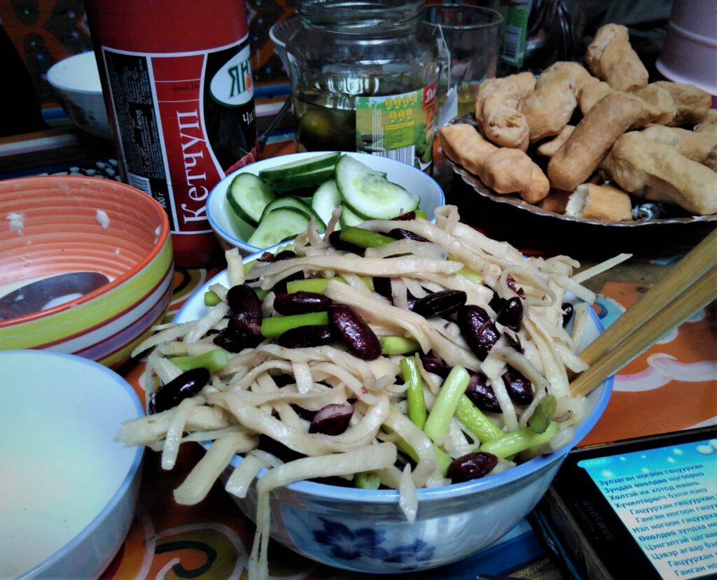Mongolische Weizennudeln mit Kidney Bohnen und Frühlingszwiebeln. Dahinter eine Schale mit Gurken, frittiertes Brot und eine Flasche Ketchup mit kyrillischer Beschriftung. 