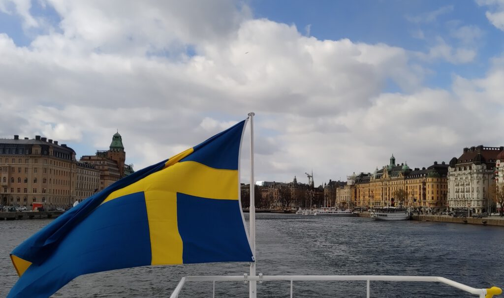 Die schwedische Fahne weht am Heck des Pendelbåts bei der Ausfahrt aus Nybroviken in der Stockholmer City. Der Himmel ist bewölkt.
