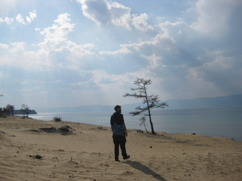 Baikal für die Seele. Ich laufe in der Mitte des Bildes auf einer Sanddüne an der Küste des Baikalsees und blicke nach rechts. Von oben Sonnenstrahlen, die die weißen Wolken durchdringen. Vor mir ein kahler kleiner Baum. Dahinter das Wasser des Baikals.