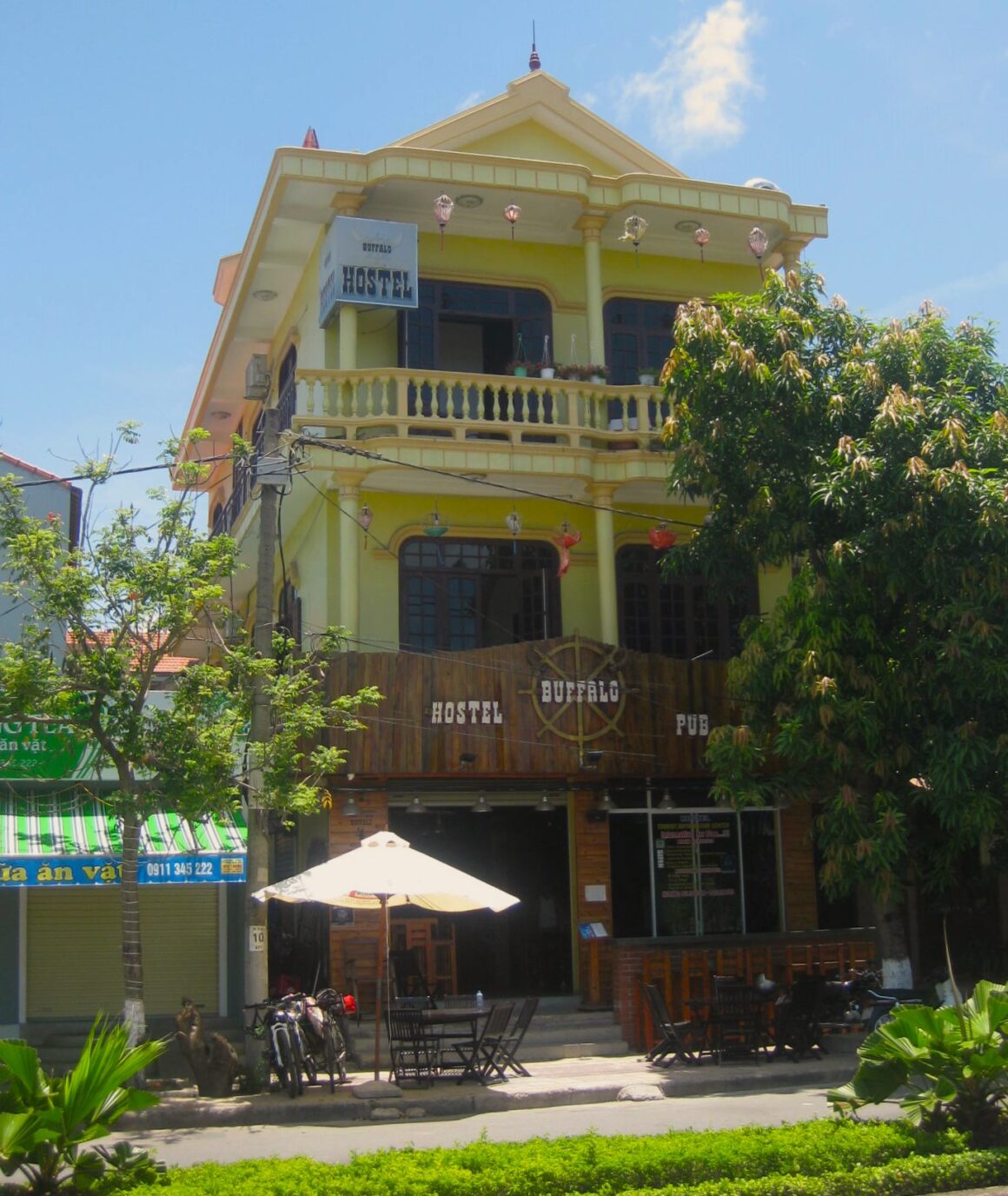 Koloniales, dreistöckiges Gebäude an einer Straße in Dong Hoi. Im Vordergrund eine Terrasse mit Regenschirm. Aufschrift auf Holz im Western-Stil "Hostel Buffalo Pub".