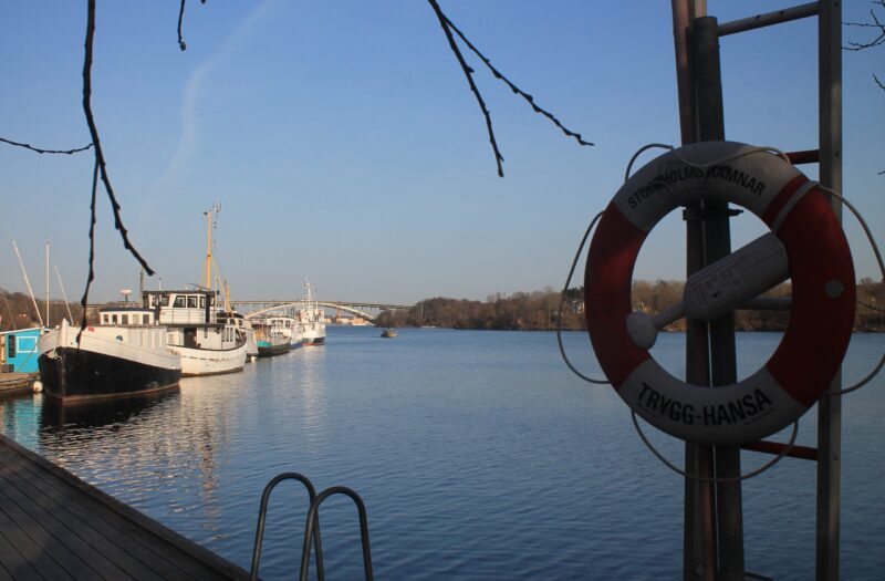 Lilla Essingen Pendelbat-Haltestelle. Rechts ein Rettungsring an einem Mast. LInks unten der Steg mit Einstiegsleiter am Wasser. Im HIntergrund liegen Boote an. Äste ragen ins Bild.