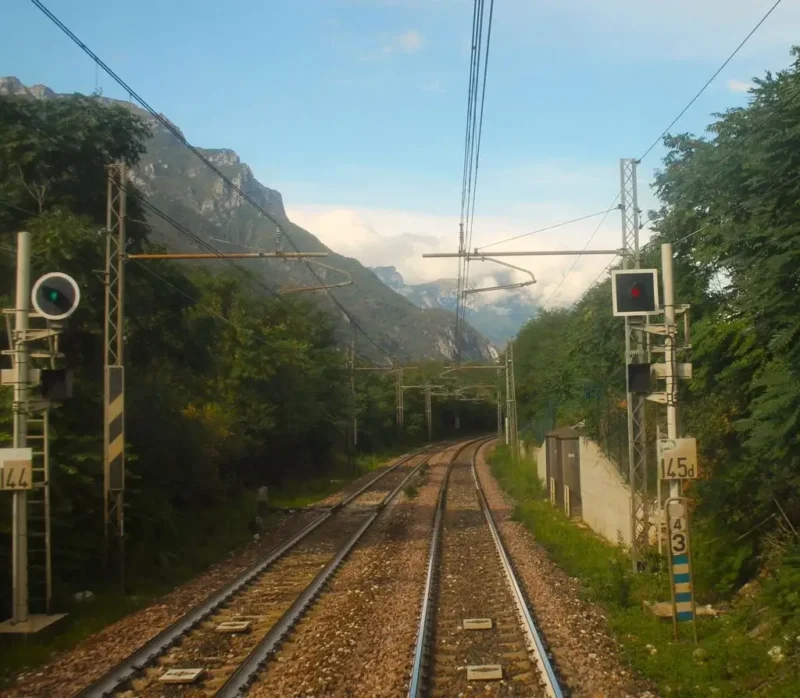 Mit dem Zug nach Venedig. Zweispurige Eisenbahnschienen zwischen den alpinen Gebirgen. Rechts rote Ampel, links grüne Ampel. Im Hintergrund schneebedeckter Berg in den Alpen.