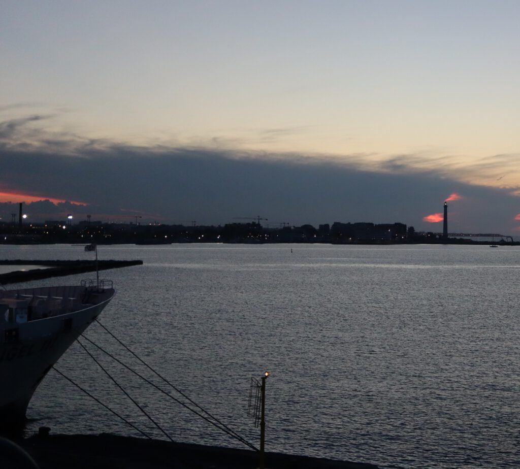 Blick über den Hafen von Bari im Dunkeln. Links ragt der Bug eines anderen Schiffes ins Bild. Rechts hinten der Leuchtturm "Bari Light" vor dem bewölkten Sonnenuntergang. 