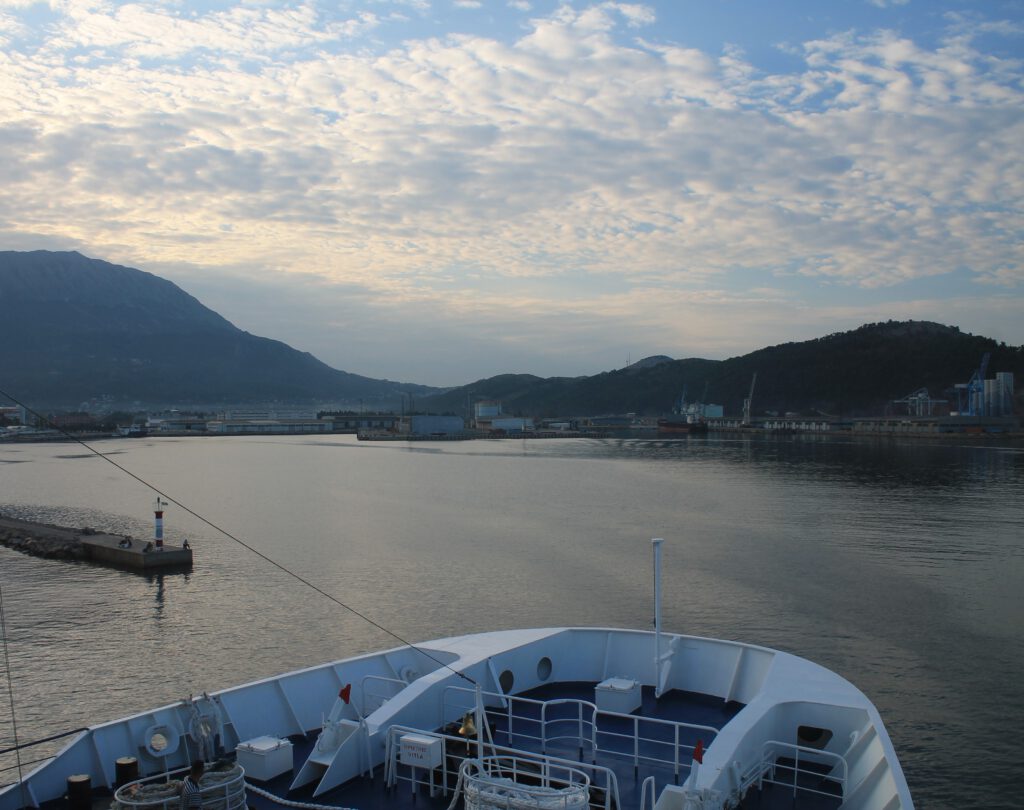 Einfahrt im Hafen von Bar Montenegro. Blick über den Hafen vom Schiff aus. Der Bug ragt ins Bild. Hinten die Gebirge vor bewölktem Morgenhimmel. Links ragt eine schmale Mole ins Hafenwasser.