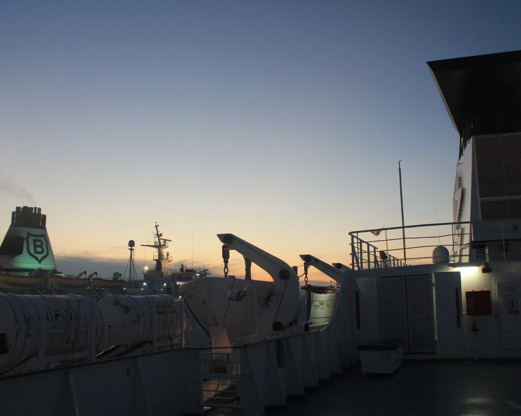 Blick über ein Außendeck der Fähre gegen den Sonnenuntergang. Links ist der Schornstein eines anderen Schiffes zu sehen. 