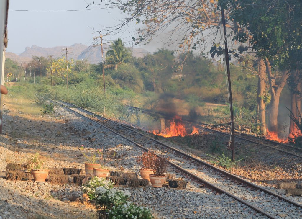 Neben dem stehenden Zug zwei Schmalspur-Schienen. Dazwischen ein frisch gelegtes Feuer mit schwarzem Rauch. Neben dem Gleis stehen 6 Blumenkübel aus Ton. 