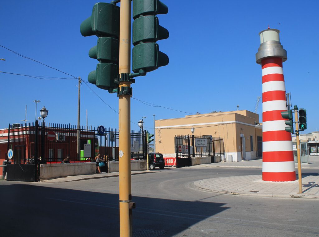 Hafen von Bari. Rechts ein kleines Modell eines rot-weiß gestreiften Leuchtturms. Links das Eingangstor. Im Vordergrund eine Ampel.