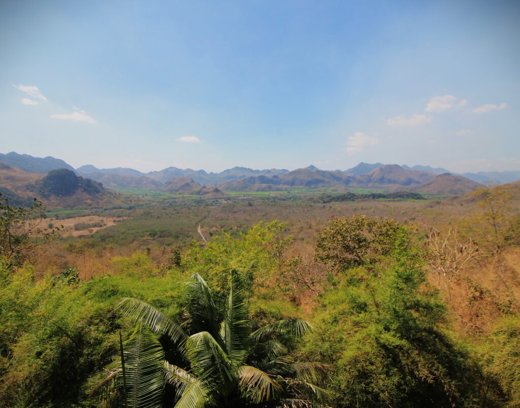 Blick über das Tal des Kwai. Im Hintergrund das Hügelland. Im Vordergrund grüne Pflanzen. Dazwischen flaches, trockenes Land. 