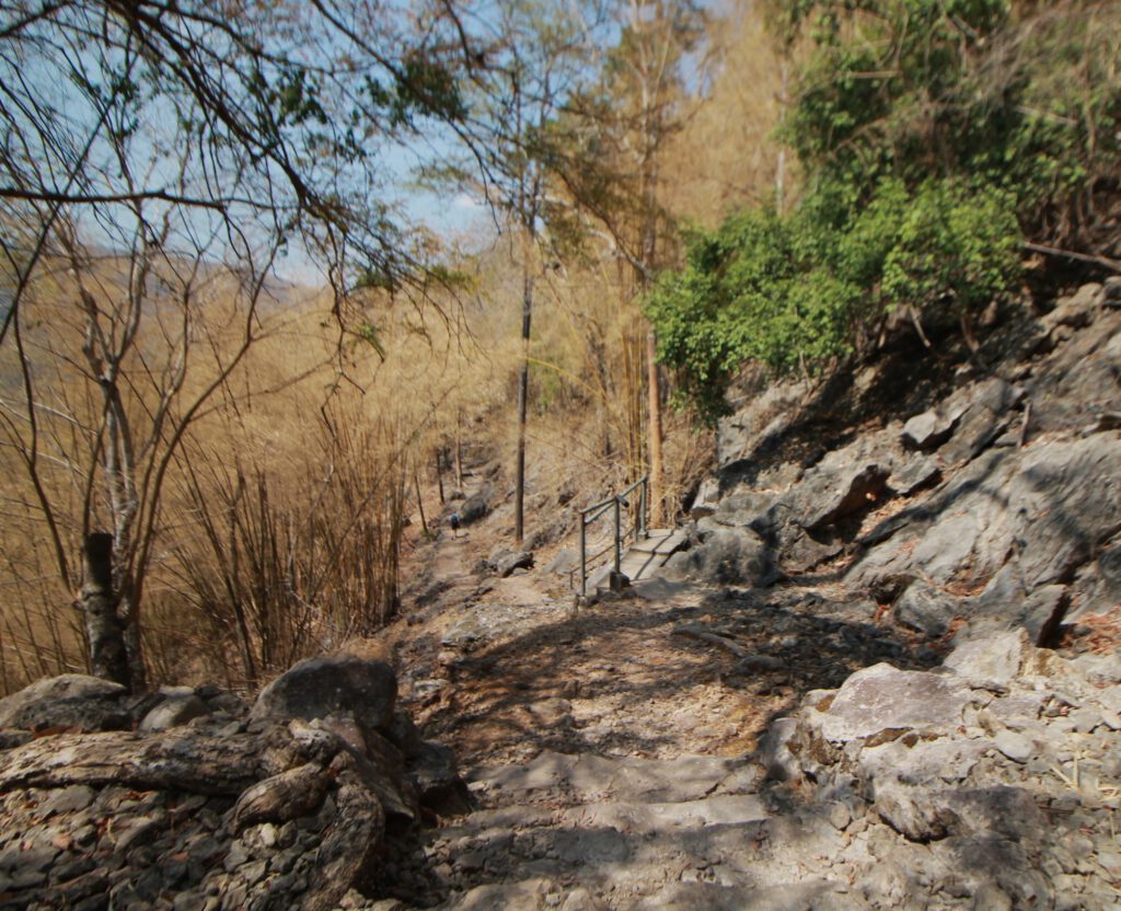 Wanderpfand entlang der ehemaligen Thailand-Burma-Eisenbahn. Der steinerne Pfad geht leicht bergab. Links trockene Pflanzen. Rechts graue Felsen.