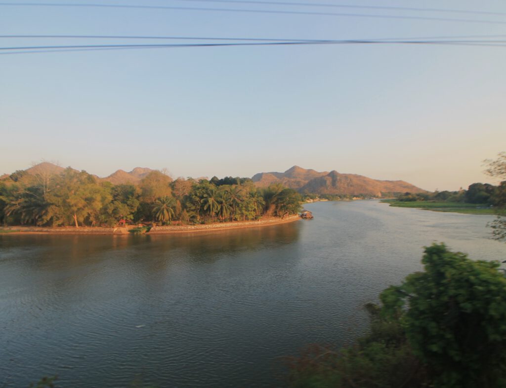 Vom Wang Pho Viadukt aus: Der River Kwai schlängelt sich S-förmig durch die trockene Hügellandschaft von Kanchanburi. Am anderen Ufer stehen Palmen und andere Bäume. 