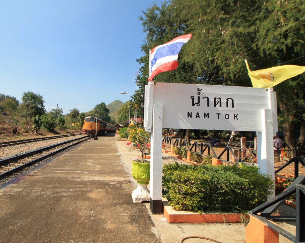 Zug der Todeseisenbahn fährt am Bahnhof in Nam Tok ein. Rechts weißes Schild mit Aufschrift "Nam Tok" in Thai-Schrift und lateinischen Buchstaben. Zwei Schienenpaare links neben dem Bahnsteig.  