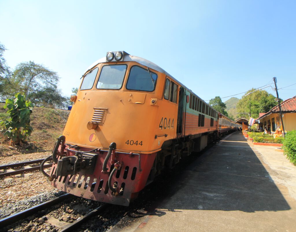 Triebwagen der Thailand-Burma-Eisenbahn: Nahaufnahme von vorne. Gelbe Front des Triebwagens erfüllt die Bildmitte. Dahinter bunte Waggons des Zugs.  