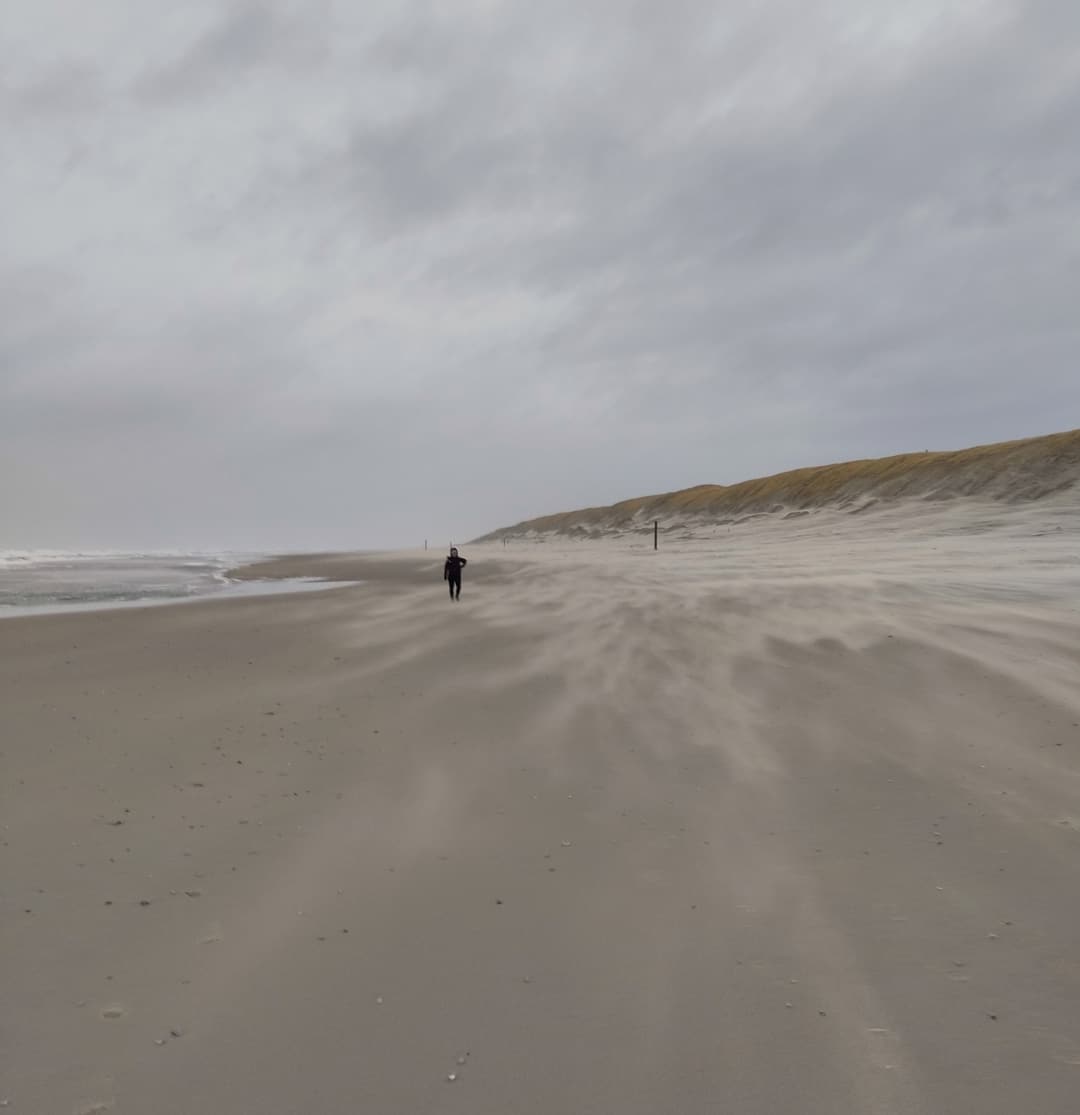 Sandsturm: Sand weht über die Oberfläche des Strandes an der Westküste von Texel. Eine Person mit Kapuze im Hintergrund. 