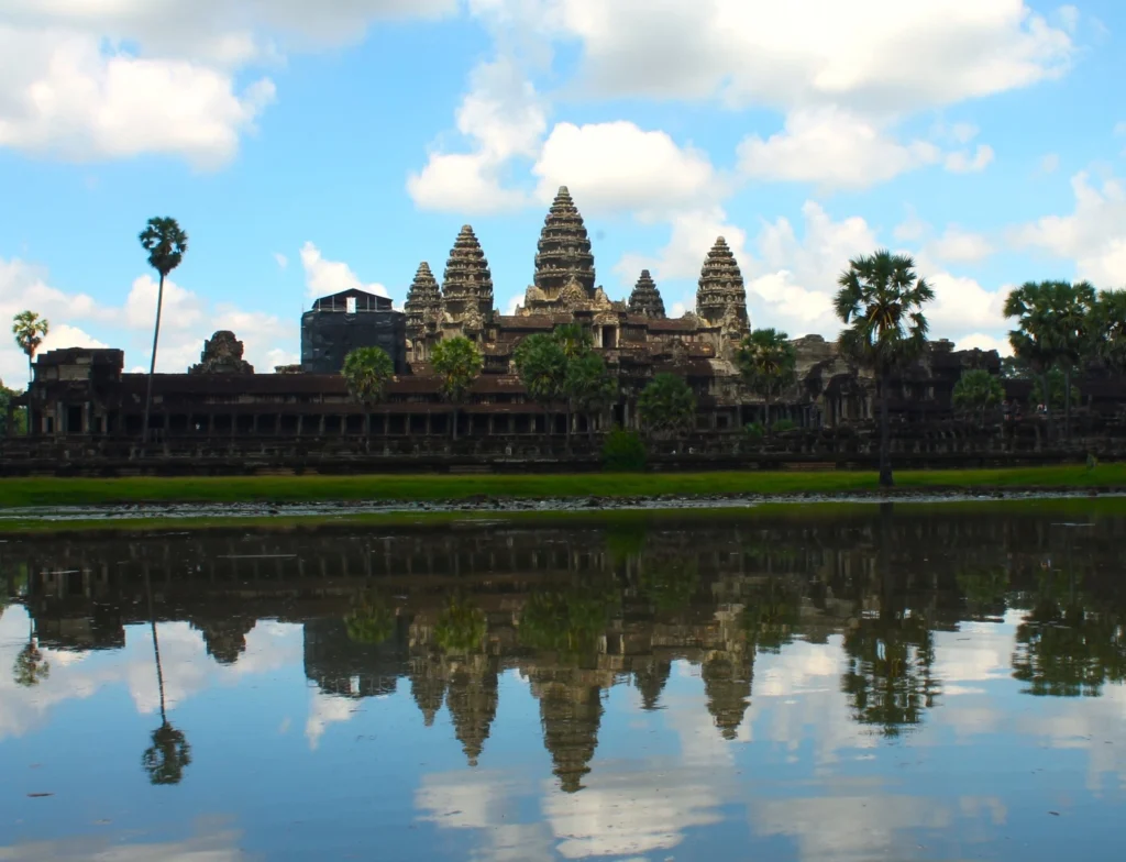 Tempel Angkor Wat, der sich im klaren Wasser des Teichs vor dem tempel spiegelt. Links im Bild ist eine Palme zu sehen, die der Szene eine tropische Note verleiht.