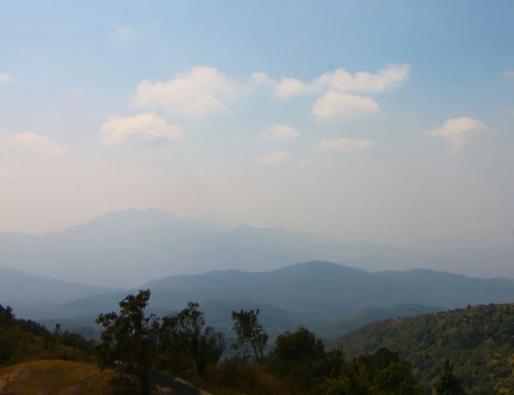 Eine Gebirgslandschaft in Nordthailand ist aufgrund von Smog in der Luft getrübt. Hügel im Hintergrund als Silhouette zu sehen. Im Vordergrund sind grüne, bewaldete Hügel sichtbar. 